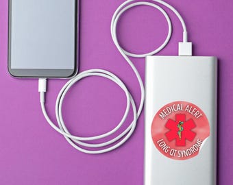 Peut inclure: Une batterie externe rectangulaire argentée reliée à un smartphone par un câble de charge blanc. La batterie porte un autocollant circulaire avec un symbole rouge de l'étoile de vie, un serpent vert et le texte MEDICAL ALERT LONG QT SYNDROME.