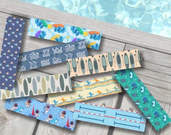 Peut inclure: Plusieurs marque-pages rectangulaires avec des motifs sur le thème de l'été. Les motifs comprennent des planches de surf, des vagues, des astronautes, des kayaks et des lignes de piscine. Les marque-pages sont disposés sur une surface en bois avec un fond d'eau.