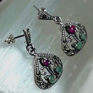 May include: A pair of silver-toned drop earrings featuring a teardrop-shaped openwork frame. Each earring is accented with one round red stone and two round green stones, complemented by small dark metallic bead-like embellishments throughout.
