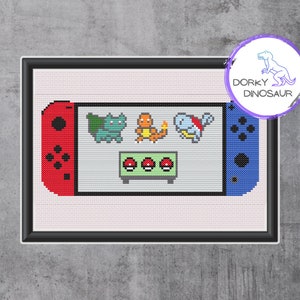 May include: Cross-stitch art of a video game console with red and blue sides. The screen displays pixelated images of three Pokémon characters and three Poké Balls. The artwork is framed and includes the text "Dorky Dinosaur".