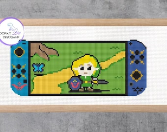May include: Cross-stitch artwork of a video game console, featuring a pixelated character in green and yellow. The console is blue and black, with a green landscape background. The artwork is framed in a light brown wooden frame. The Dorky Dinosaur logo is in the upper left corner.