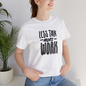 Puede incluir: Camiseta blanca con texto negro que dice "Less Talk -More- Work" en una fuente en negrita y estilizada.