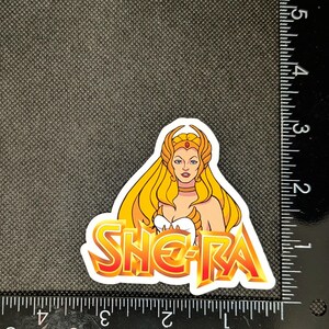 May include: A sticker featuring She-Ra, with long blonde hair, a golden crown, and a white and gold outfit. The sticker has the word "SHE-RA" in a fiery orange and yellow font. The sticker is approximately 3 inches in height.