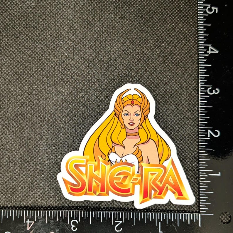 May include: A sticker featuring She-Ra, with long blonde hair, a golden crown, and a white and gold outfit. The sticker has the word "SHE-RA" in a fiery orange and yellow font. The sticker is approximately 3 inches in height.