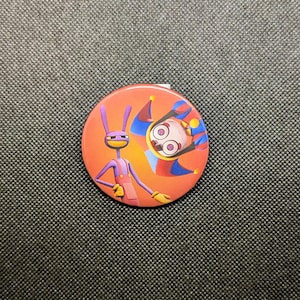 May include: A round, orange button featuring two cartoon characters. One is a purple figure with a yellow smile and overalls. The other is a jester with a white face and large eyes, wearing a blue, red, and yellow hat.