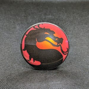 May include: A round, black and red pop socket featuring a dragon silhouette. The dragon is black, set against a gradient of red and orange. The pop socket is a gaming accessory.