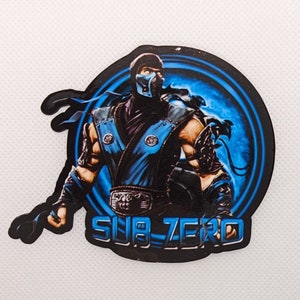 May include: A black and blue sticker featuring a character from the video game Mortal Kombat. The character is Sub-Zero, a ninja with a blue mask and a blue and black outfit. The sticker has a blue and white background with a circle around the character. The text "SUB-ZERO" is written in white letters on a black background.