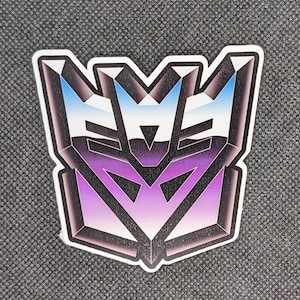 May include: A purple, blue, and white sticker with a geometric design resembling the Autobot symbol. The sticker is shaped like a crest or shield.