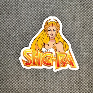 May include: A sticker featuring a cartoon illustration of She-Ra with long blonde hair, a golden crown, and a white top. The word "SHE-RA" is in a bold, orange and yellow font.