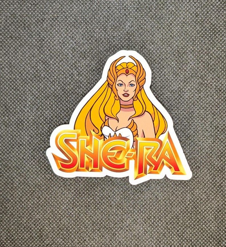 May include: A sticker featuring a cartoon illustration of She-Ra with long blonde hair, a golden crown, and a white top. The word "SHE-RA" is in a bold, orange and yellow font.