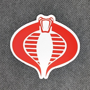 May include: Red and white Cobra logo sticker. The Cobra is a stylized snake with a red body and white stripes. The Cobra's head is facing forward with its mouth open.