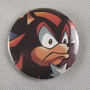 May include: A round button featuring the character Shadow the Hedgehog from the Sonic the Hedgehog video game series. The button has a black background with red, orange, and white accents, and a stern expression.