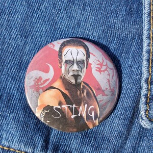 May include: A round button with a picture of Sting, a professional wrestler, wearing a black and white face paint. The button has a red background with white flowers and the word "STING" in white letters.