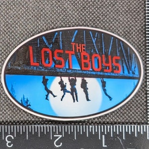 K&ouml;nnte beinhalten: Ein blauer und wei&szlig;er ovaler Aufkleber mit dem Text "The Lost Boys" in roten Buchstaben. Der Aufkleber zeigt die Silhouette von f&uuml;nf Figuren, die an einem Holzbalken h&auml;ngen.