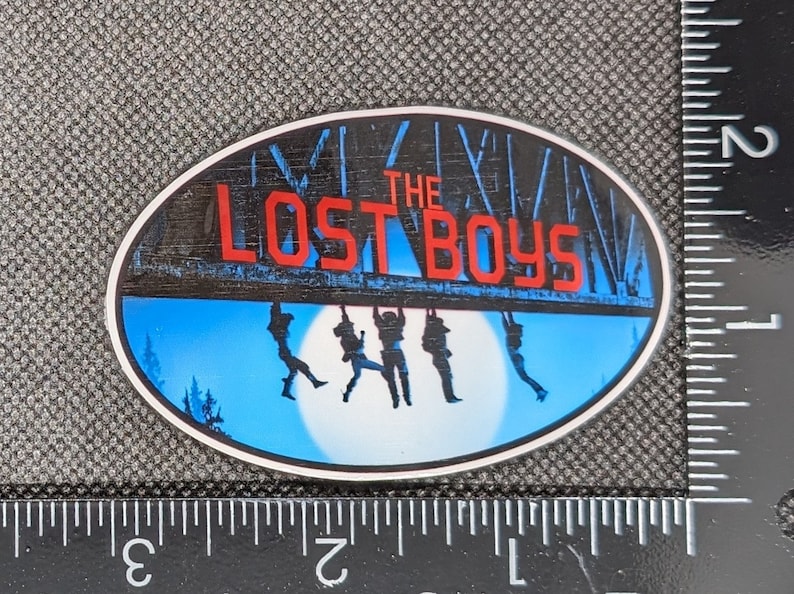 K&ouml;nnte beinhalten: Ein blauer und wei&szlig;er ovaler Aufkleber mit dem Text "The Lost Boys" in roten Buchstaben. Der Aufkleber zeigt die Silhouette von f&uuml;nf Figuren, die an einem Holzbalken h&auml;ngen.