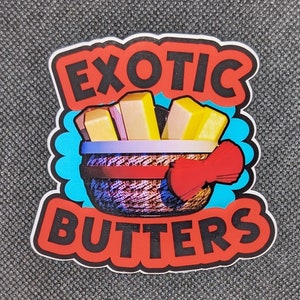 May include: A red and blue sticker with the text "Exotic Butters" in black letters. The sticker features a cartoon image of a basket with a red bow and yellow sticks of butter inside.