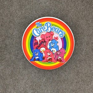 May include: A round sticker with a rainbow border and the text "The Care Bears" in a white circle. The sticker features a group of six Care Bears, including a pink bear with a heart, a blue bear with a star, a yellow bear with a sun, a green bear with a rainbow, a purple bear with a moon, and a brown bear with a wishbone.