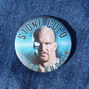 May include: A round button with a blue background and a picture of Steve Austin, also known as Stone Cold Steve Austin, a professional wrestler. The text "Stone Cold Steve Austin" is written around the edge of the button in a black font.
