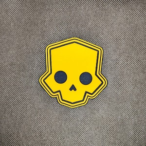 May include: A yellow skull-shaped patch with a black outline and two dark blue eye sockets and a nose. The patch is on a gray textured surface.