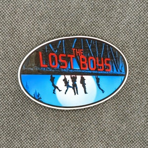 K&ouml;nnte beinhalten: Ein runder Aufkleber mit blauem und wei&szlig;em Hintergrund, der eine Szene mit vier Figuren zeigt, die an einer Holzbr&uuml;cke h&auml;ngen. Die Br&uuml;cke tr&auml;gt den Text "THE LOST BOYS" in roten Buchstaben.