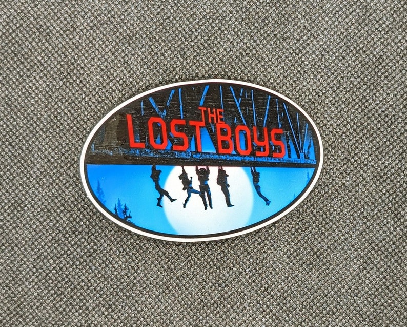K&ouml;nnte beinhalten: Ein runder Aufkleber mit blauem und wei&szlig;em Hintergrund, der eine Szene mit vier Figuren zeigt, die an einer Holzbr&uuml;cke h&auml;ngen. Die Br&uuml;cke tr&auml;gt den Text "THE LOST BOYS" in roten Buchstaben.