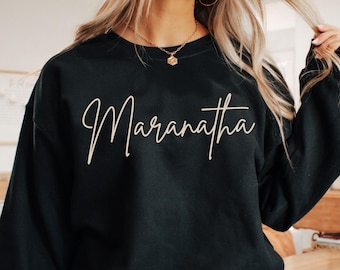 Könnte beinhalten: Schwarzes Sweatshirt mit dem Wort "Maranatha" in Schreibschrift. Das Sweatshirt hat lange Ärmel und einen Rundhalsausschnitt. Das Wort "black" befindet sich in der unteren rechten Ecke des Bildes.
