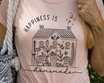 May include: A light peach-colored t-shirt with the words "HAPPINESS IS homemade" printed in black. The graphic features a house with floral details. The shirt is knotted at the waist and paired with a tote bag.