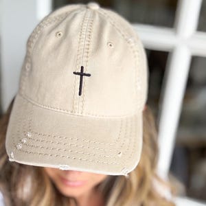 May include: A beige baseball cap with a distressed look and a small black embroidered cross on the front.