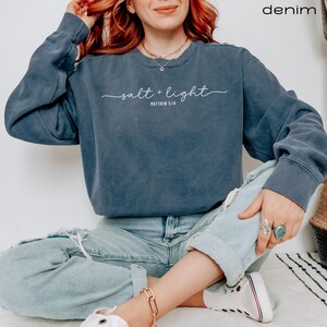 Puede incluir: Una sudadera azul oscuro con el texto "salt + light" y "Matthew 5:14" en letras blancas. La modelo lleva la sudadera con pantalones vaqueros azules.