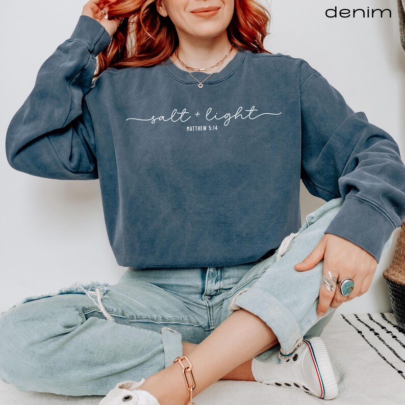 Puede incluir: Una sudadera azul oscuro con el texto "salt + light" y "Matthew 5:14" en letras blancas. La modelo lleva la sudadera con pantalones vaqueros azules.