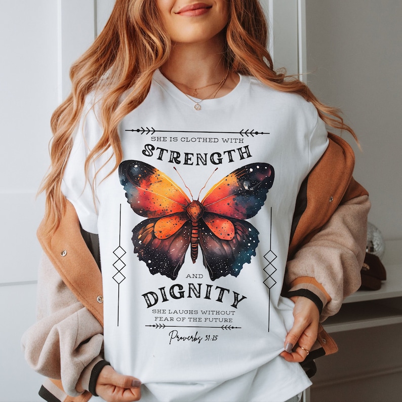May include: White t-shirt featuring a colorful butterfly design with the words "She is clothed with Strength and Dignity" and the Bible verse Proverbs 31:25. The butterfly has orange, red, and blue hues.