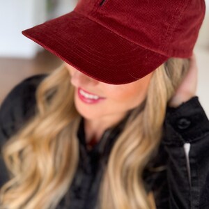 May include: A burgundy corduroy baseball cap with a black embroidered cross on the front.