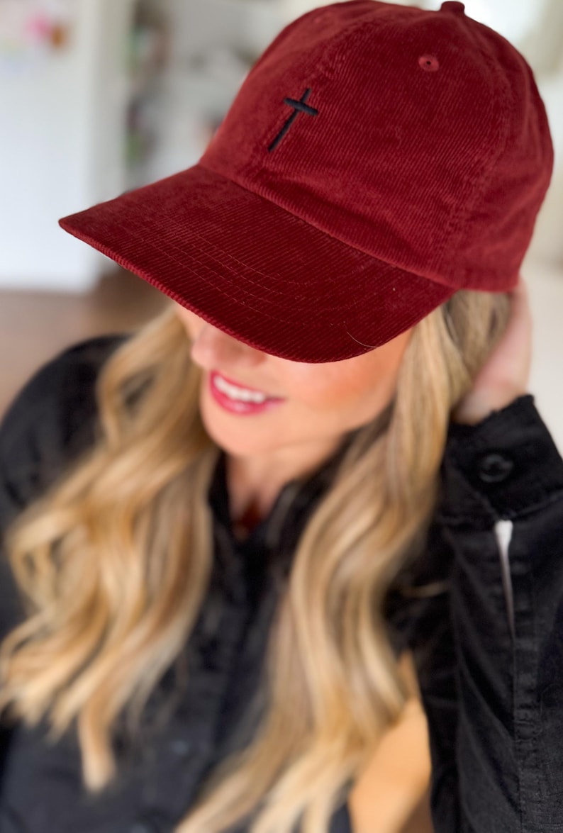 May include: A burgundy corduroy baseball cap with a black embroidered cross on the front.
