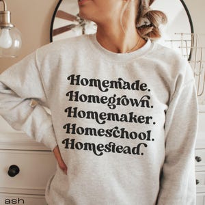 Op de afbeelding: Een grijs sweatshirt met de tekst "Homemade. Homegrown. Homemaker. Homeschool. Homestead." gedrukt in zwart.
