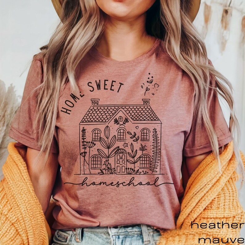 Pode incluir: Uma camiseta cor malva claro com um gr&aacute;fico preto de uma casa com o texto "Home Sweet Homeschool" e um design floral.