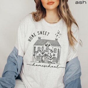 Pode incluir: Uma camiseta branca com um gr&aacute;fico preto de uma casa com flores e o texto "Home Sweet Homeschool".