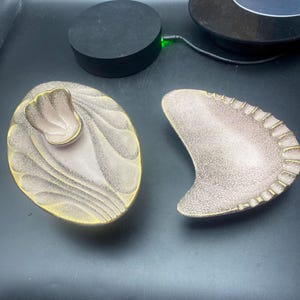 May include: Two vintage ashtrays. One is oval with a pink and gold design, and the other is a kidney shape with a similar pink and gold design. Both have a textured surface and are likely made of ceramic.