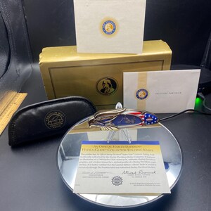 May include: A black and gold Harley-Davidson Hydra-Glide Collector Folding Knife with a red, white, and blue American flag design. The knife is displayed on a black circular stand with a mirrored surface. The knife is accompanied by a certificate of authenticity signed by Stuart Pankratz, Vice President of Harley-Davidson Motor Company.