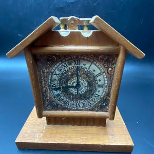 May include: A wooden clock shaped like a house with a gold-colored face and Roman numerals. The clock has a small, hinged roof that opens to reveal a lighter.