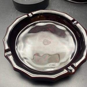 May include: A burgundy-colored ashtray with a scalloped edge and four cigarette rests. The ashtray has a glossy finish and a circular interior. The ashtray is made of a solid material.