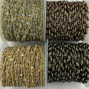 Könnte beinhalten: Vier Spulen mit Perlenketten in verschiedenen Farben: Grau, Dunkelrot, Beige und Schwarz. Jede Kette besteht aus kleinen, facettierten Perlen, die mit goldfarbenem Draht aufgefädelt sind. Die Spulen befinden sich auf einer weißen Oberfläche.
