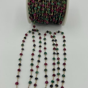 May include: A spool of beaded chain with alternating ruby, emerald, and black beads. The beads are strung on a gold-colored chain. Several strands of the beaded chain are unspooled, displaying the pattern.