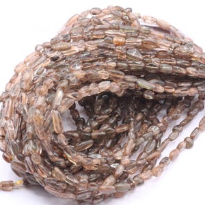 May include: A strand of brown and clear faceted gemstone beads. The beads are long and narrow, resembling small, elongated pebbles.