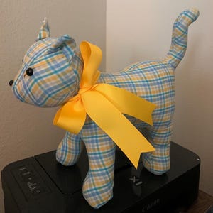 May include: A handmade stuffed cat in a plaid pattern of yellow, blue, and white. The cat has black button eyes and a bright yellow ribbon tied around its neck. The cat is standing on a black printer.