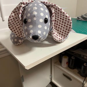 May include: A handmade plush dog toy with a gray body and white polka dots. The dog has large floppy ears with a floral pattern and a white bow. The toy has black button eyes and a black nose.