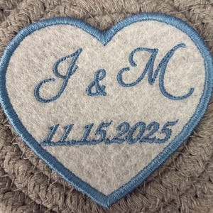 May include: A heart-shaped patch with a light blue border and embroidered text. The text includes the letters "J & M" above the date "11.15.2025". The patch is set against a gray, woven background.