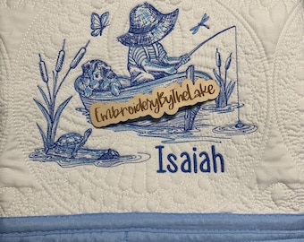 May include: A white quilted baby blanket with blue embroidery of a boy in a boat fishing. The boat has a sign that says "Embroidery by the Lake". The blanket has a blue border and the name "Isaiah" is embroidered below the image.