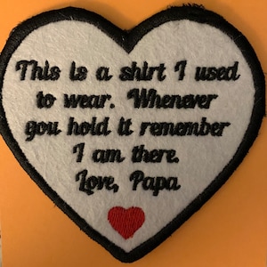 May include: White felt heart-shaped patch with black embroidered stitching around the edges. The patch has black embroidered text that reads "This is a shirt I used to wear. Whenever you hold it remember I am there. Love, Papa". A small red felt heart is embroidered in the bottom center of the patch.