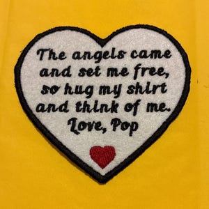 May include: Heart-shaped patch with black trim and the text "The angels came and set me free, so hug my shirt and think of me. Love, Pop." A small red heart is at the bottom. The background is yellow.