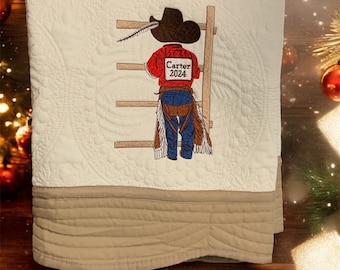 Peut inclure: Couverture crème avec une bordure beige. La couverture présente un motif brodé d'un cow-boy sur une échelle, portant un chapeau et une chemise rouge avec "Carter 2024". La couverture a un motif matelassé.